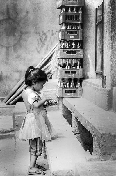 Little Girl With A Ponytail Standing In Front Of A General Store (Nepal)